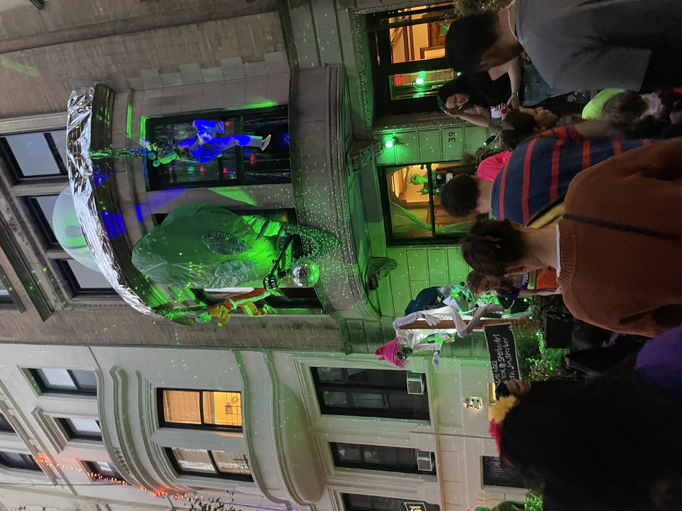 Trick-or-treaters gather outside a brownstone with a glowing green alien and flying saucer Halloween display